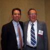 White man in black suit jacket and tie standing next to older white man in glasses and gray suit and tie