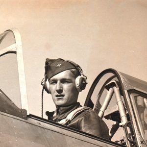 White pilot in open cockpit wearing military uniform with cap and headphones