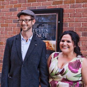 White man smiling in glasses and hat standing next to white woman smiling in dress with poster on brick wall behind them