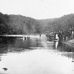 Crowd of people gathered at a tree-lined river watching a baptism