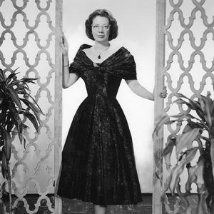 White woman with glasses in dress between decorative panels and plants