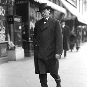 White man in great coat and hat, walking down city sidewalk