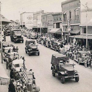 Military vehicles on town street with crowds of people and parked cars on both sides