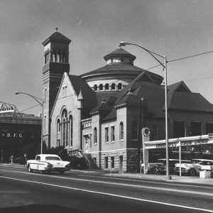 Elaborate religious building with tall bell tower and round dome on city street with a used car lot next to it