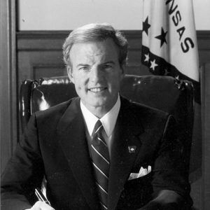 White man in suit and tie smiling at his desk with Arkansas flag behind him