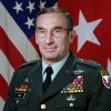 White man with glasses in military uniform with flags behind him