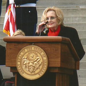 White woman with glasses and black coat speaking at lectern