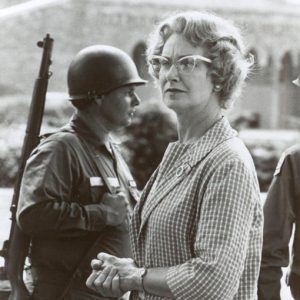 White woman with glasses in checkered shirt standing with white soldiers in uniform with guns outside school building