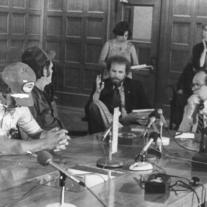 White men talking at table with microphones and white women standing behind them