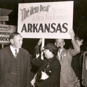 white man and woman talking in front of other white people holding political signs including "The New Deal Never a Step Backward Arkansas"