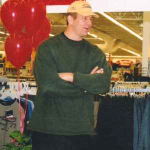 White man in sweater and cap standing next to balloons inside store