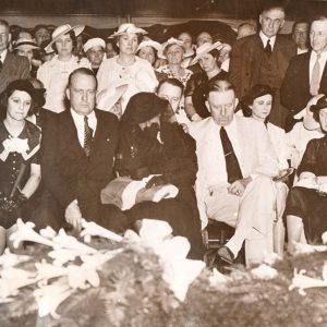 Crowd of white men and women seated and standing before flower-draped casket