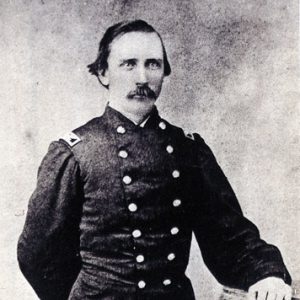 dark-haired white man with mustache in military uniform poses with left hand resting on a column