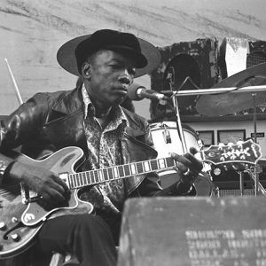 African-American man wearing black hat and playing electric guitar on stage with drums behind him and speaker in foreground