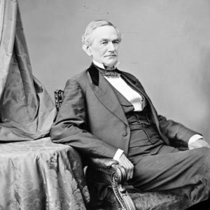 Older white man in suit high collar and bow tie posing in a chair with his elbow on a table covered in ornate cloth