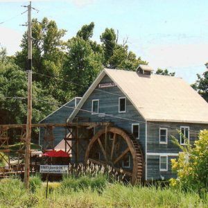 Three-story mill building with water wheel and aqueduct with more buildings and trees in the background