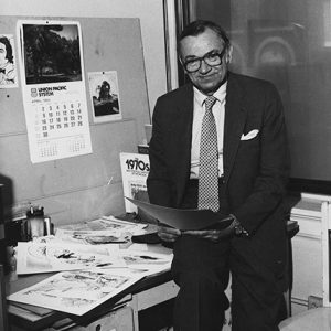 Older white man with glasses in suit sitting on desk in office