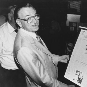 Older white man with glasses in suit smiling as he holds a page from the Arkansas Democrat about him in his hands