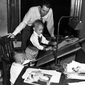 White man standing and looking over two white boys at desk wtih art supplies