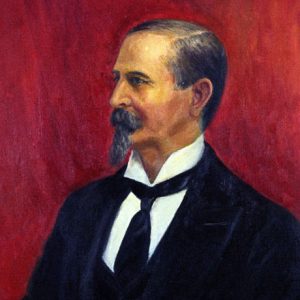 Profile view of white man with mustache and long chin beard in suit before a red background