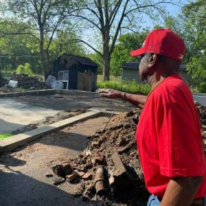 African-American man in red shirt and hat standing next to pile of rubble pointing towards new foundations under construction