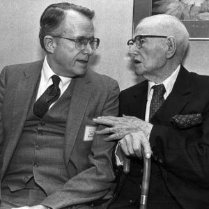 Two older white men in suits and glasses seated conversing with the elder gesturing holding cane