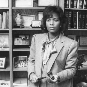 black woman in suit and silk necktie with gavel standing in front of bookshelf