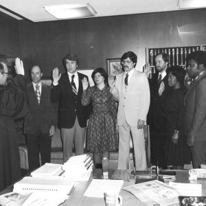 White man in black robes holding up his right hand in front of white men in suits and ties and and a woman in a dress who are also holding up their right hands