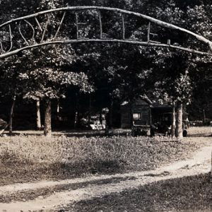 White children at both sides of camp entrance gate with arch above on dirt road