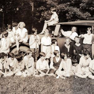 children and adults mostly dressed in white gathered around a car with an older man behind the wheel