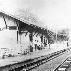 group of men and women at on platform of depot building with steam locomotive coming in on tracks