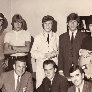 Young white men, one with a guitar, standing and white men in suits sitting in front of them