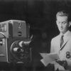 White man in suit and tie reading papers next to television camera