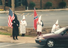 White men wearing Ku Klux Klan outfits and carrying confederate and American flags on street corner