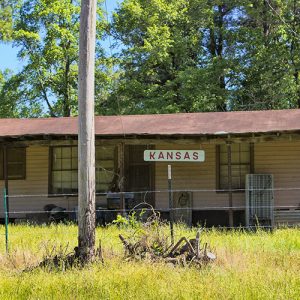 Single-story house inside barbed wire fence with hanging "Kansas" sign