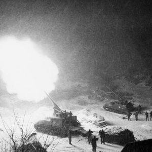 Two artillery cannons, one of them firing into the air, with soldiers covering their ears amid heavy snow