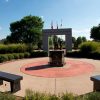 Round concrete platform with two benches and statue in center and concrete entrance way with three flag poles