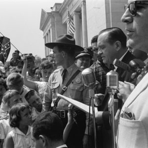 White man in suit speaking into microphones outside classical stone building with guard and crowd with american flag and confederate flag