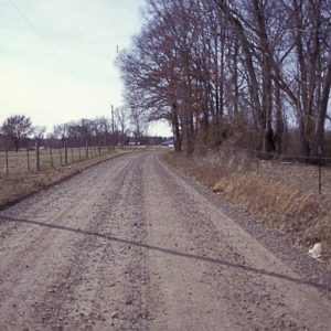 gravel road with barbed wire fence and field on the left and trees on the right