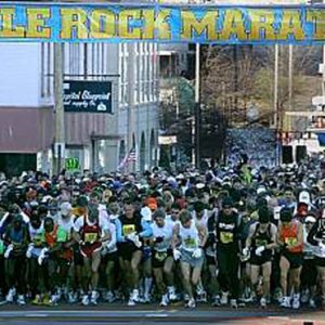 Mixed crowd of people in shorts standing on city street with "Little Rock Marathon" banner hanging above them