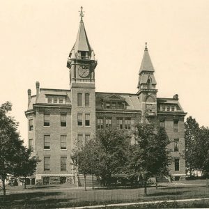 Multistory brick building with clock tower and second tower with trees in its front yard