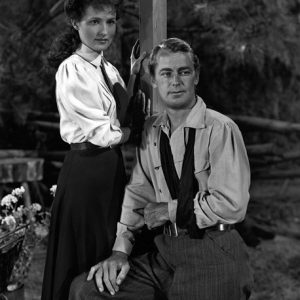 Young white woman with pulled-back hair standing next to young white man sitting with his left arm in a sling