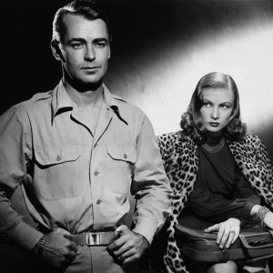 White man standing next to white woman sitting in leopard print jacket with suitcase