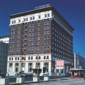 Tall hotel building on street corner with "LaFayette" sign on top next to a paid parking lot with red trimmed booth