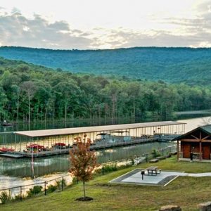 small wooden building next to lake with floating covered dock with parked boats, tree-covered hills in background