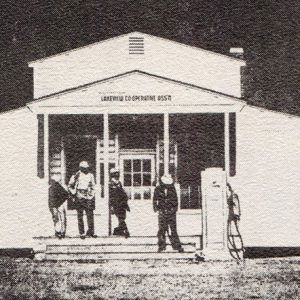 Four men standing on steps of building with combination roof and gas pump in front