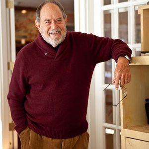 Older white man in red sweater with his left arm resting on a shelf