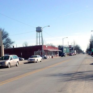 Town street lined with buildings and parked cars with trees and water tower in the background