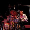 Older white man in white shirt and black pants smiling behind drum set on stage