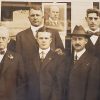 Group of white men in suits standing with posters in window behind them